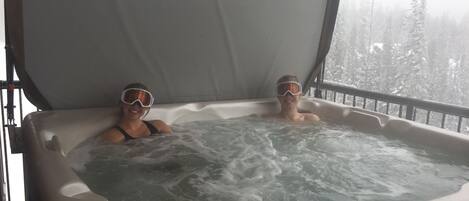 Un bain à remous où deux personnes portent des lunettes de protection, entouré d’une terrasse en bois et offrant une vue sur une forêt enneigée.