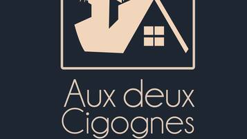 En logo med et stiliseret hus og et træ, og teksten 'Aux deux Cigognes'.