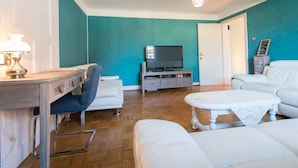 A living room with a television, a desk with a lamp, a sofa, and a small table. - Appartement aux Deux Cigognes (Gries)