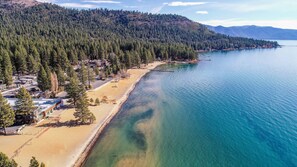 A coastal area with a sandy beach, a clear blue lake, and a forested hillside.
