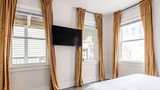 A hotel room with a bed, a flat-screen TV mounted on the wall, and large windows with curtains.