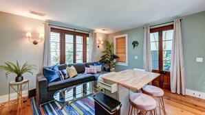 A modern living room with a sofa, a dining table, and a coffee table. There are two windows with curtains, a potted plant, and a lamp on the wall. - Monaco Manor | Charleston Home w/ Rooftop Deck! (Charleston)