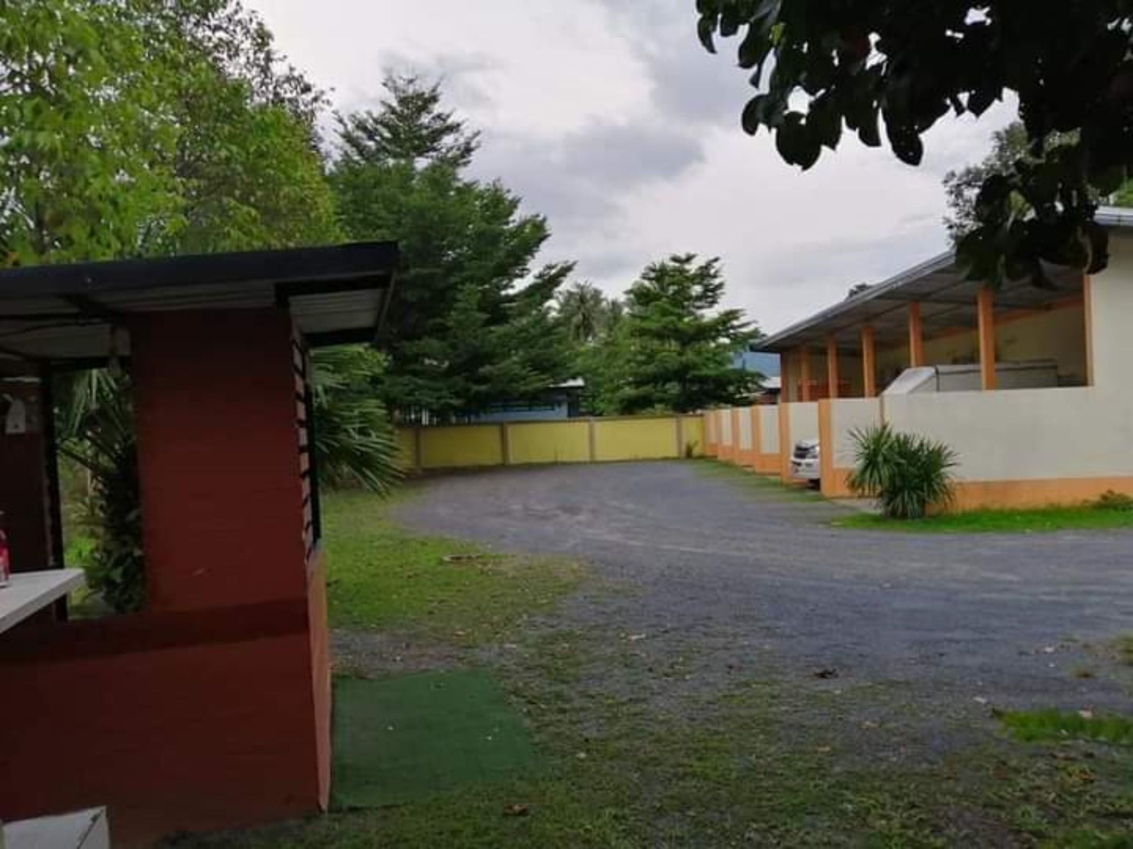 A building with a covered outdoor area, a paved driveway, and a small garden with a tree.
