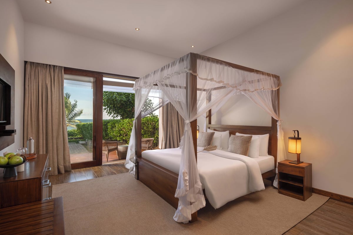 A four-poster canopy bed with a nightstand and lamp, a television, and a dining area with a fruit bowl.