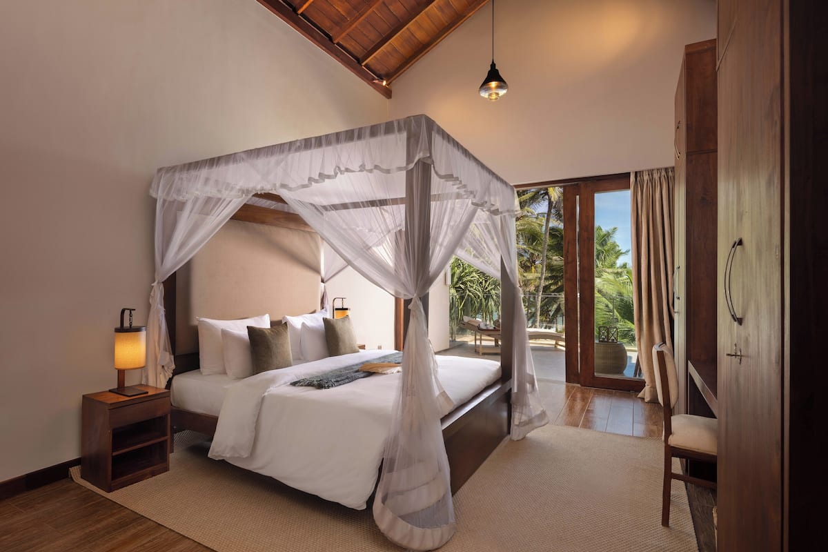 A four-poster canopy bed with a nightstand, a lamp, and a view of the outdoors.