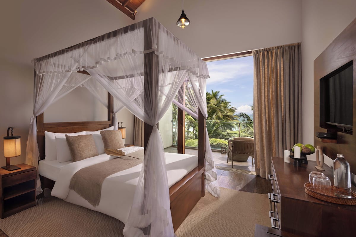 A spacious bedroom with a four-poster bed, a bedside table with a lamp, and a view of palm trees through the window.
