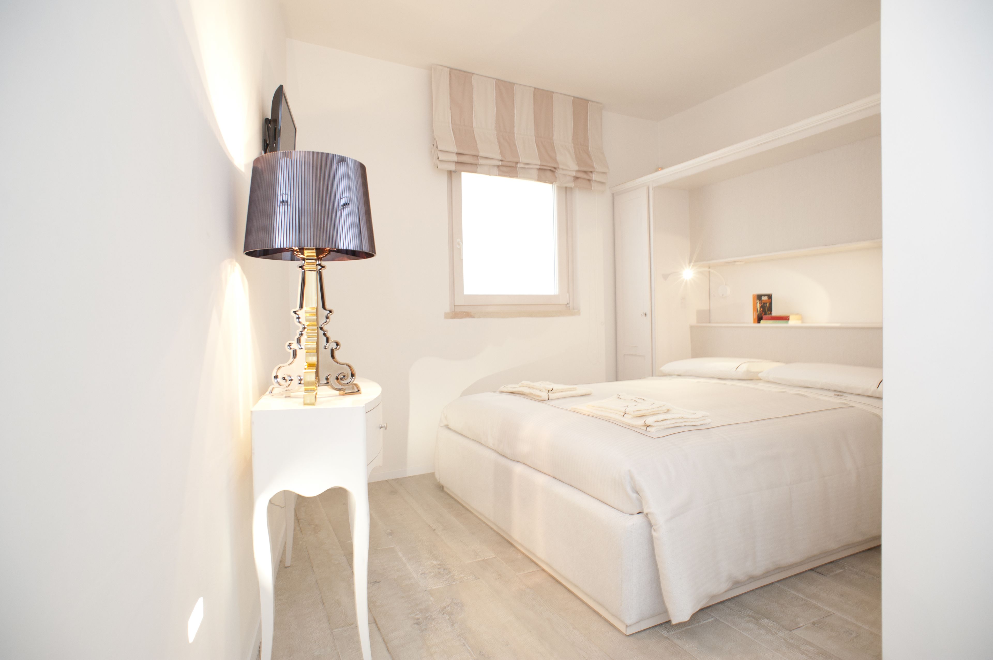 A bedroom with a bed, a bedside table with a lamp, a window with curtains, and a shelf with books.