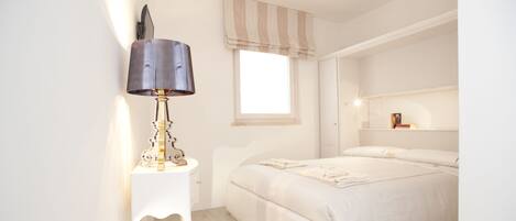 A bedroom with a bed, a bedside table with a lamp, a window with curtains, and a shelf with books.