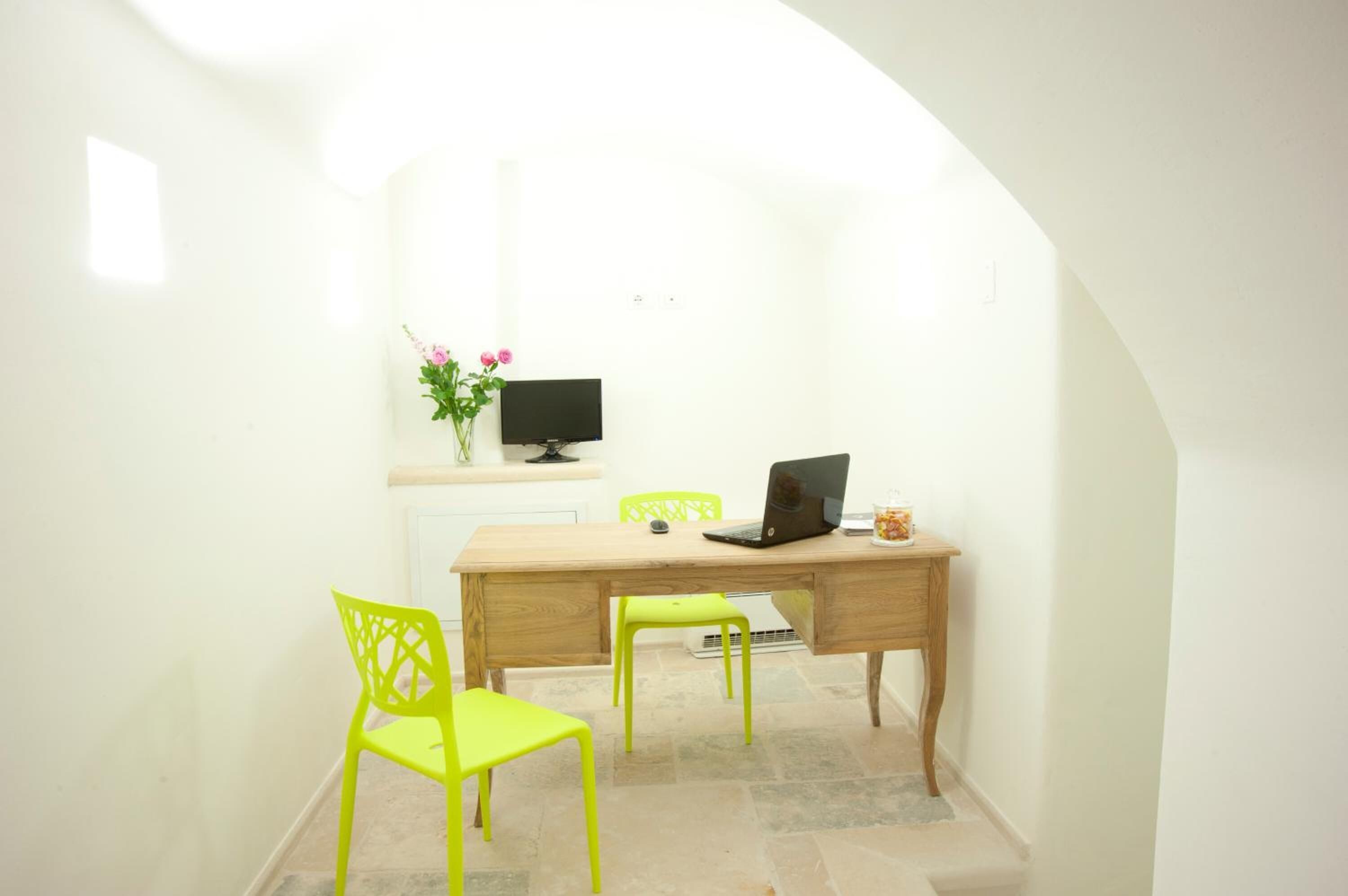 A small, bright room with a wooden desk, two green chairs, a laptop, a TV, and a vase with flowers.