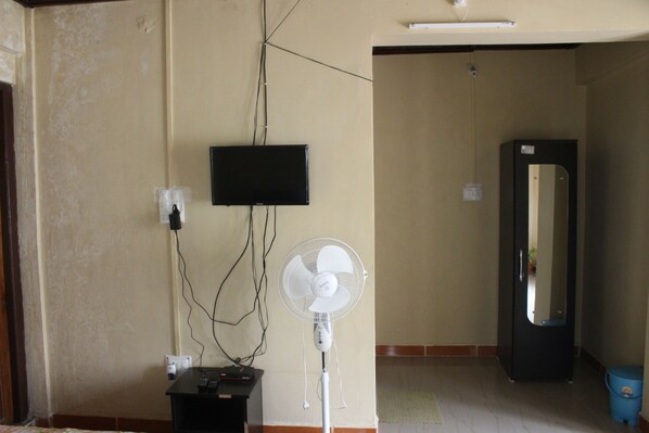 A room with a flat-screen TV mounted on the wall, a standing fan, and a door leading to another room.