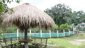 A thatched-roof structure with a bench underneath, surrounded by greenery and a fence. - Pumpkin StreamSide Guest House (Shillong)