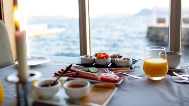 A breakfast table with various dishes and a glass of orange juice, set against a view of the sea. - Stad Hotell (Stad)