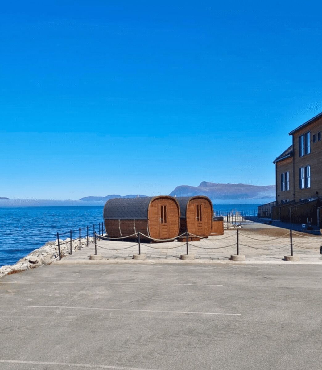 Une promenade en bord de mer agrémentée de barils en bois, un sentier pavé et un bâtiment doté de grandes fenêtres.