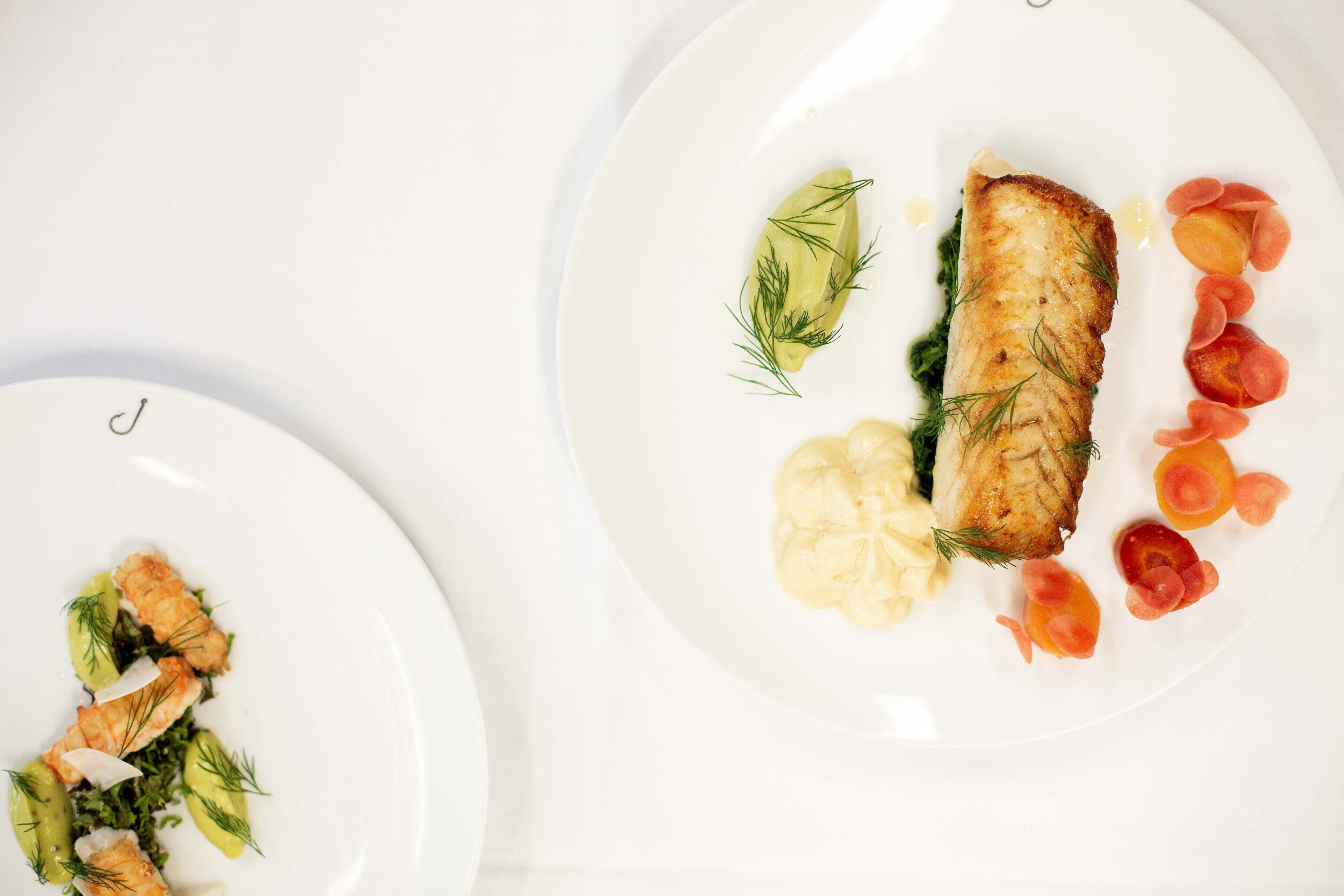 Two white plates with food, one with a fish and vegetables, the other with a fish and a creamy side.