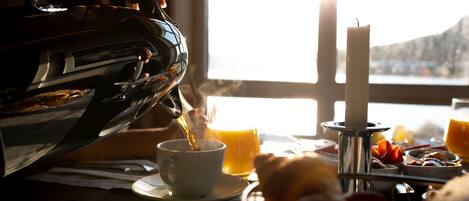 Una escena de desayuno con un hervidor sirviendo té en una taza, una vela y una mesa con diversas comidas y un vaso de jugo de naranja.