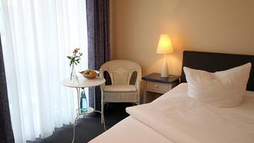 A hotel room with a bed, a nightstand, a lamp, a chair, a small table with a vase of flowers, and a window with curtains.