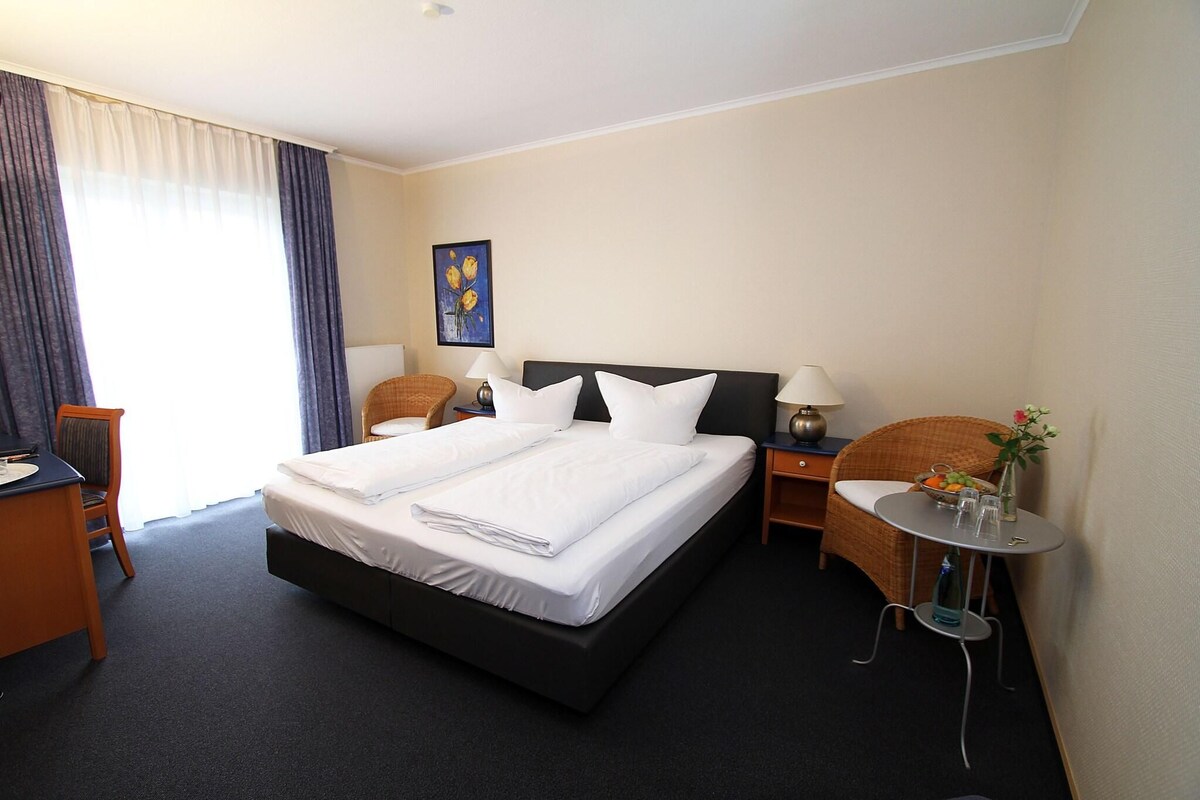 A hotel room with a large bed, two bedside tables, a chair, a small table with a vase, and a painting on the wall.