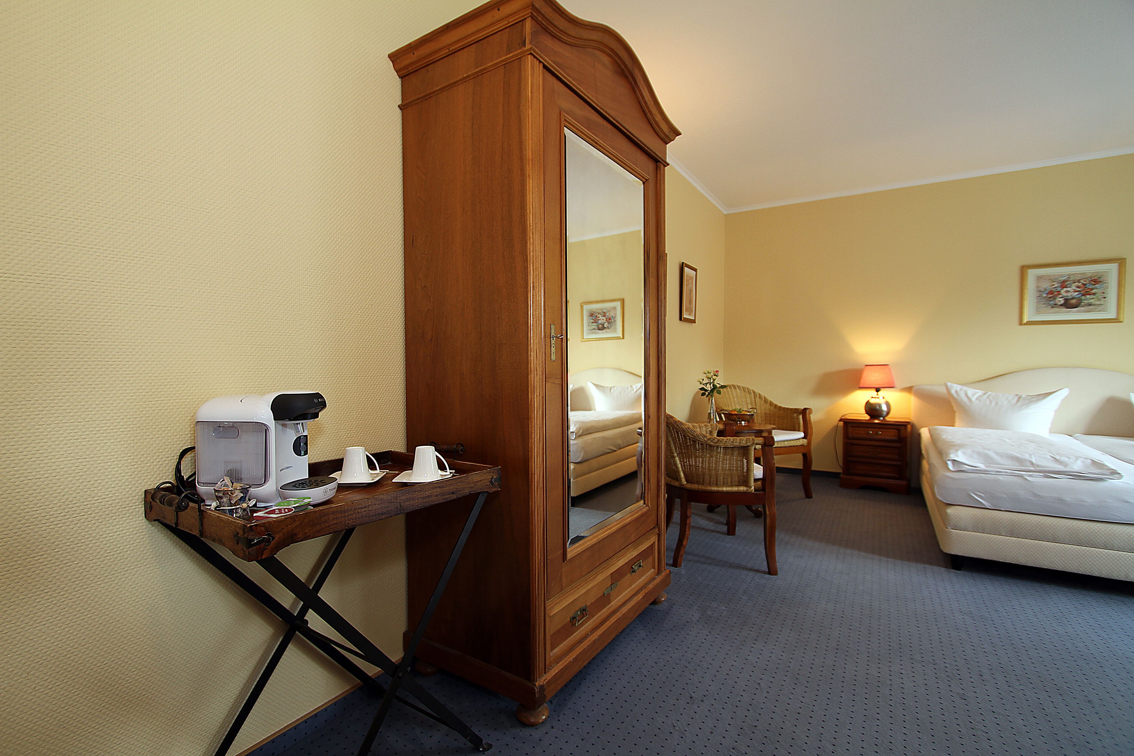 A hotel room with a wooden wardrobe, a small table with a coffee maker, a chair, a bedside table with a lamp, and a bed.