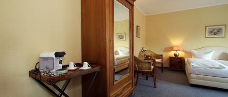 A hotel room with a wooden wardrobe, a small table with a coffee maker, a chair, a bedside table with a lamp, and a bed.