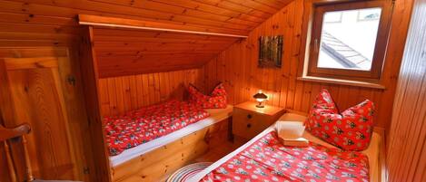 Una habitación de cabaña acogedora con paredes de madera y techo inclinado. Hay dos camas con ropa de cama y almohadas rojas a juego, una mesita de noche con una lámpara y una ventana que proporciona luz natural.
