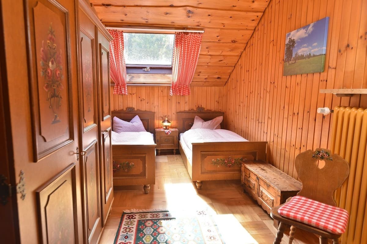 Una habitación acogedora con paredes y suelos de madera, dos camas con cabeceros a juego, una mesita, una silla, una ventana con cortinas y un radiador.