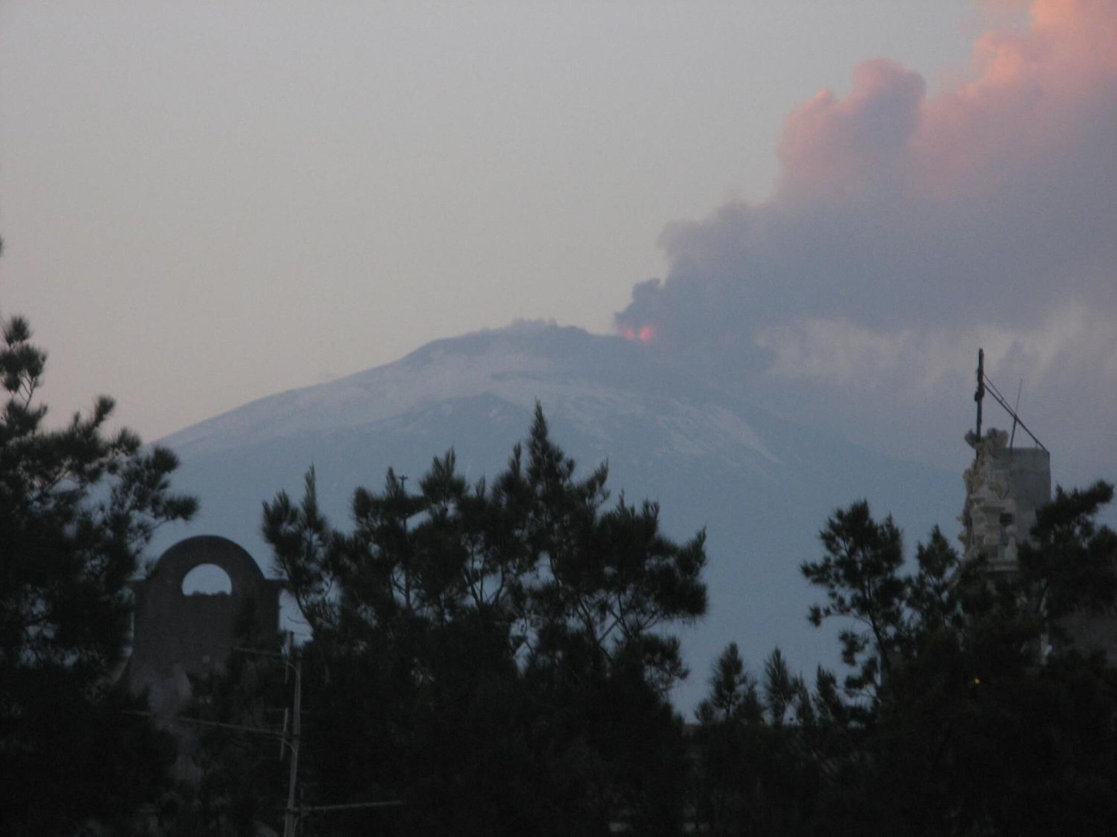 Una montaña con una columna de humo visible, rodeada de árboles y una estructura con una abertura circular.