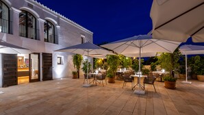 Une terrasse extérieure avec des tables et des chaises, entourée de plantes en pot et d’ombrelles.