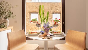 Un coin repas avec une table dressée pour deux personnes, offrant une vue sur un mur de pierre extérieur à travers une fenêtre.