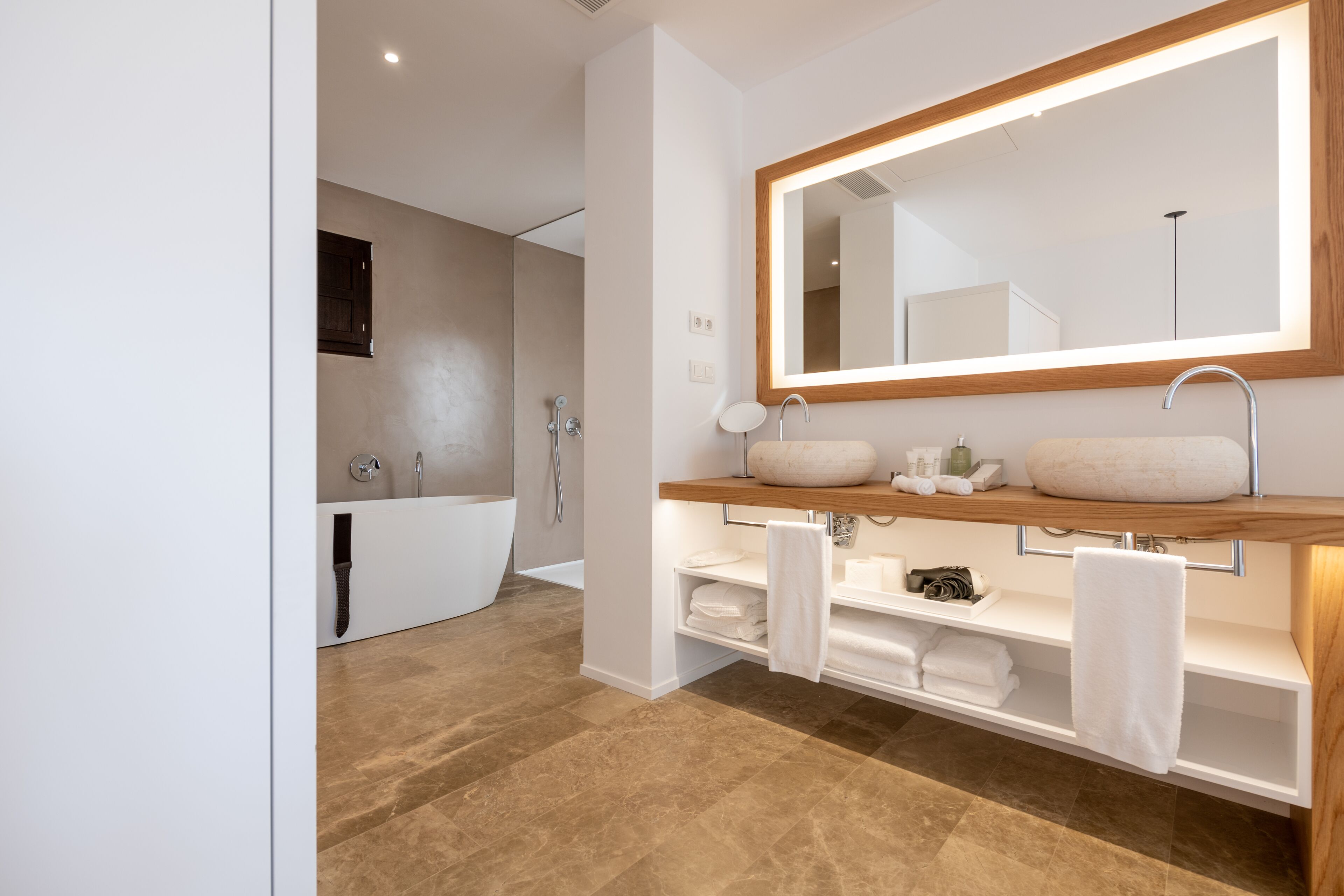 A modern bathroom with a large mirror, two sinks, a bathtub, and a vanity with shelves for towels.