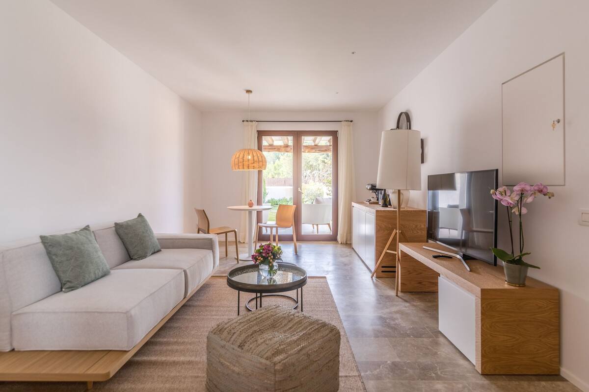 Een moderne woonkamer met een bank, voetenbankje, salontafel en televisie. Er is een eethoek met stoelen en een tafel, en een keuken met een aanrecht en kasten.