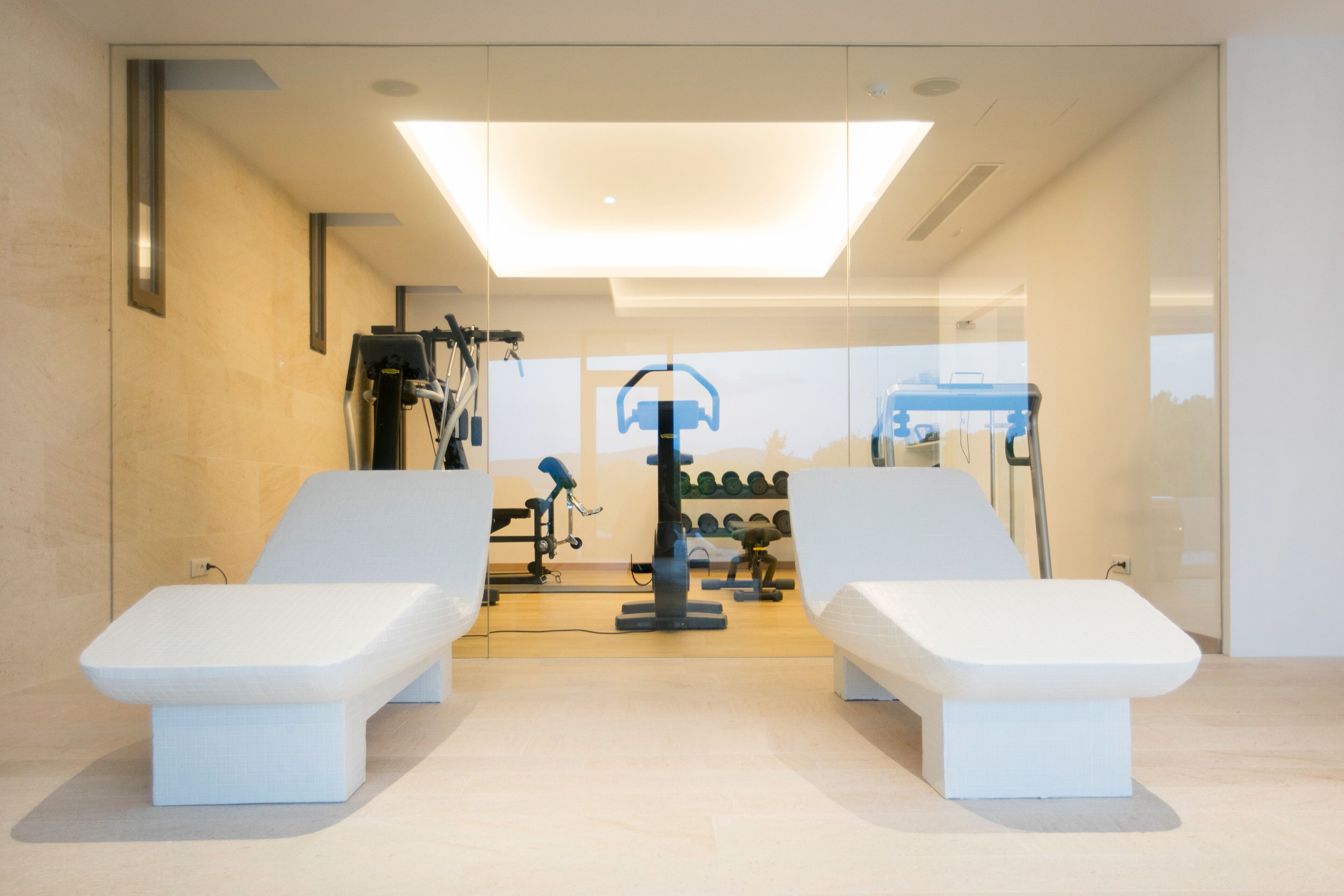 Una habitación moderna y bien iluminada con dos mesas de masaje blancas, una máquina de ejercicios montada en el techo y un estante de mancuernas fijado a la pared.