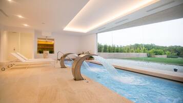 A modern indoor pool with a waterfall feature, lounge chairs, and a view of the outdoors.
