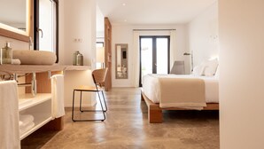Une chambre d’hôtel moderne dotée d’un grand lit, d’un coin salon avec un fauteuil, d’une coiffeuse avec un miroir et d’une salle de bain comprenant un lavabo et un porte-serviettes.
