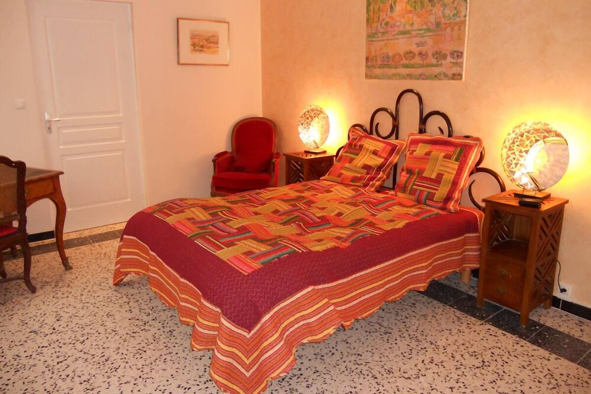 Une chambre à coucher avec un grand lit, deux lampes de chevet, un fauteuil rouge, une porte et un tableau encadré au mur.