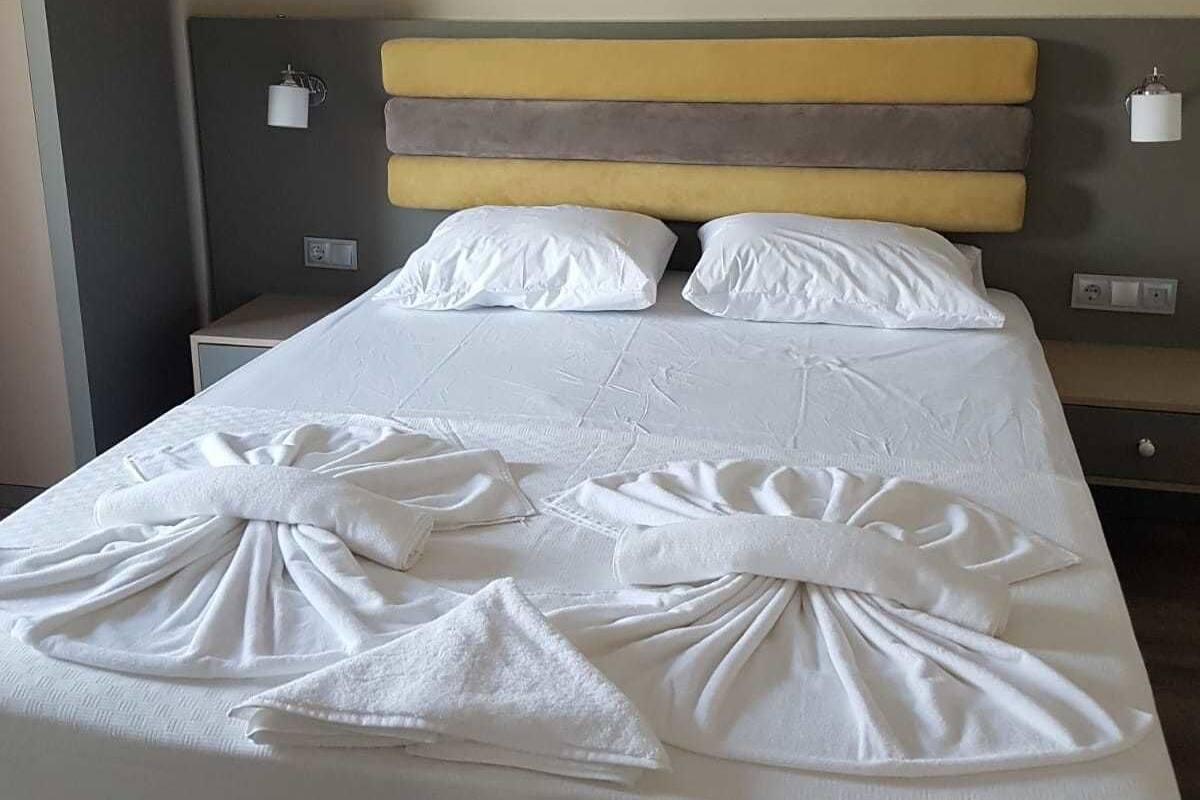 Una cama bien hecha con sábanas y toallas blancas dispuestas en forma de lazo, cabecero con rayas horizontales, mesitas de noche con lámparas, y una lámpara de techo roja.