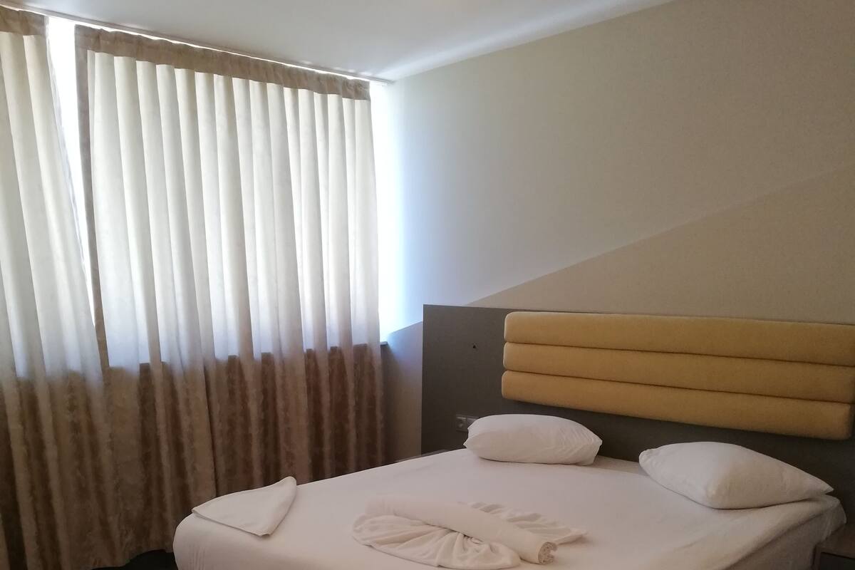 Una habitación de hotel con una cama, cabecera y una ventana con cortinas.