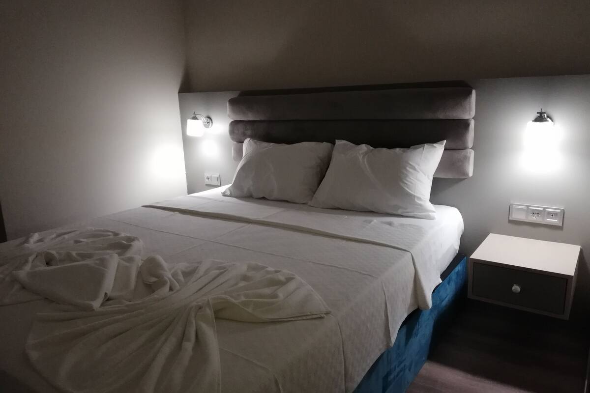 Una cama bien hecha con edredón blanco y almohadas, una mesita de noche con una lámpara y un enchufe de pared.