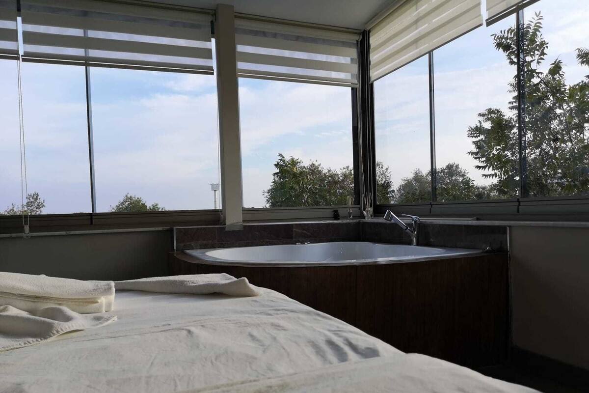 Un baño moderno con una bañera grande, una ventana con persianas y vistas a árboles.