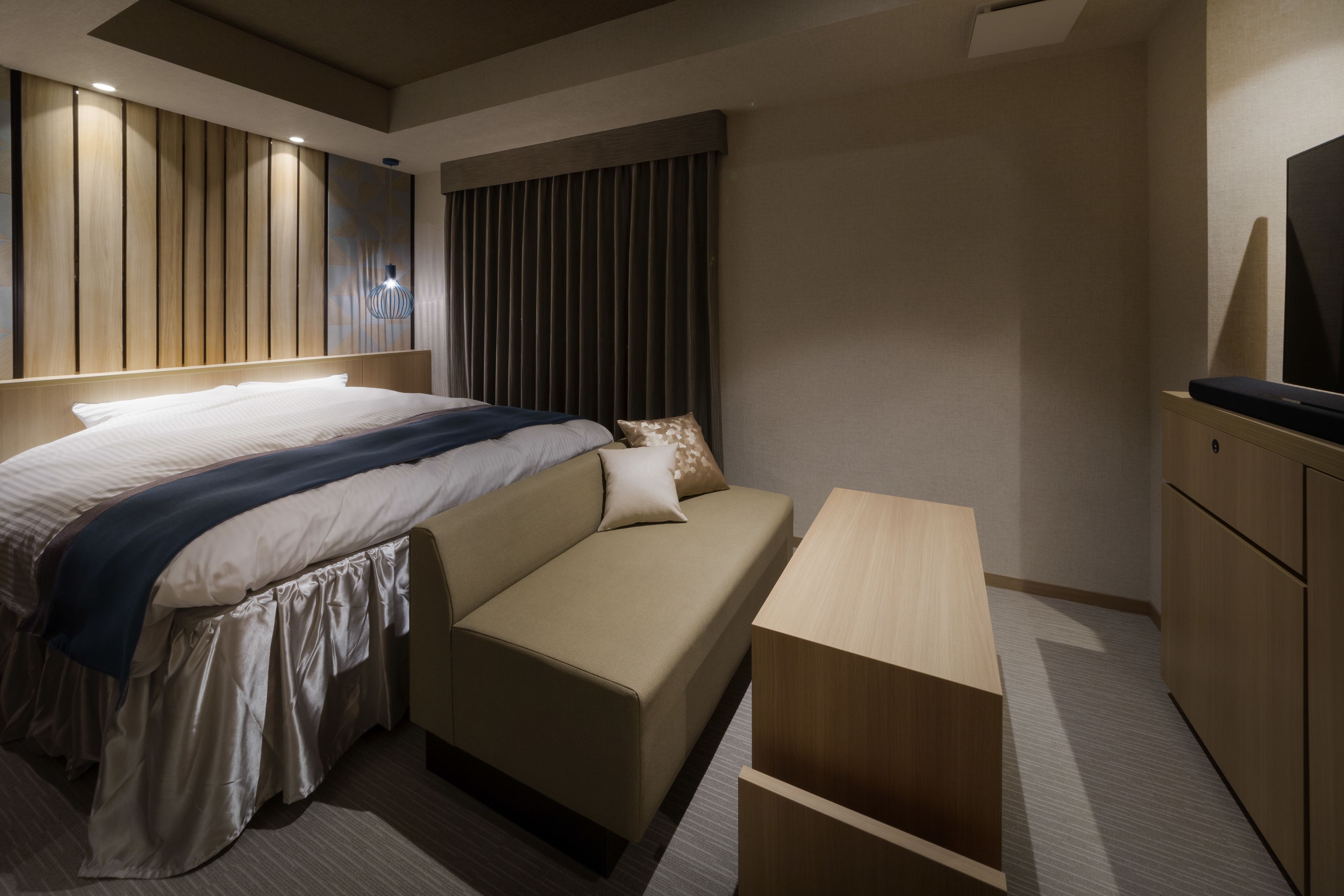 A modern hotel room with a large bed, a sofa, a wooden nightstand, and a TV.