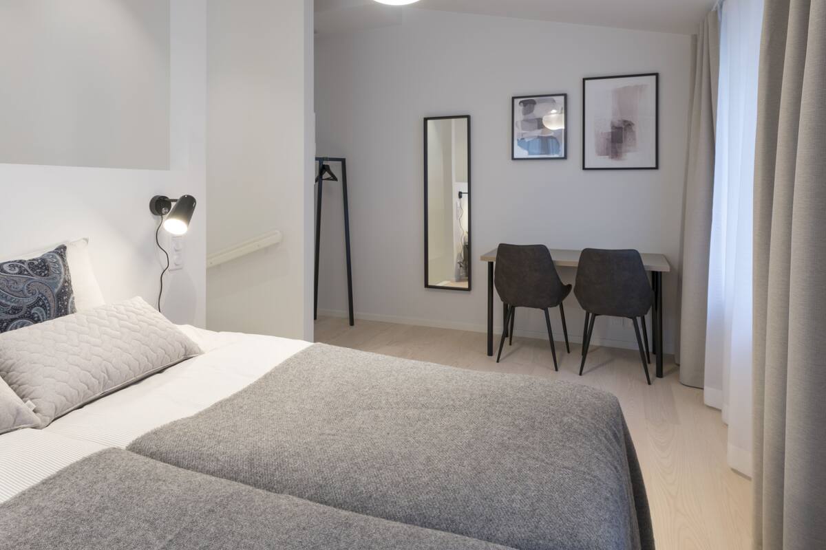 Quarto de hotel moderno com uma cama grande, duas cadeiras, uma escrivaninha, um espelho e uma luminária fixada na parede.