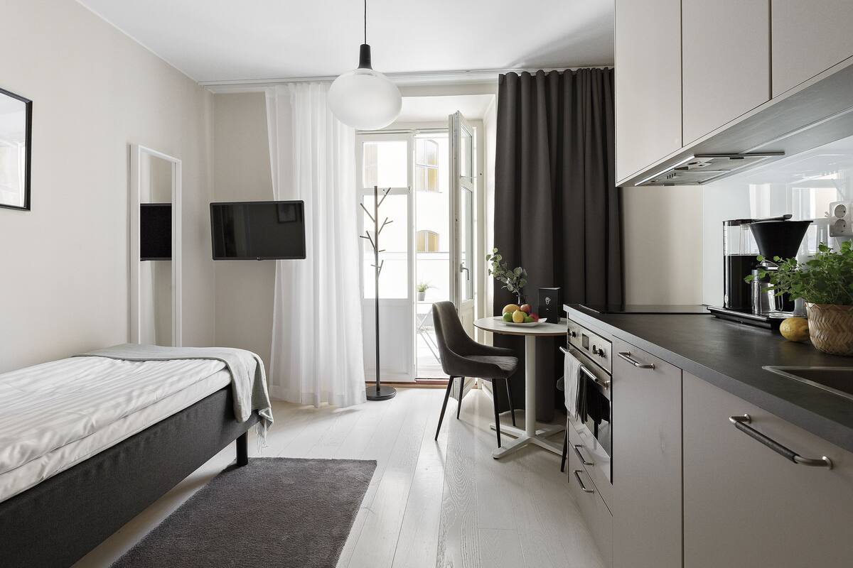Quarto de hotel moderno com cama, escrivaninha, cozinha compacta e uma grande janela.
