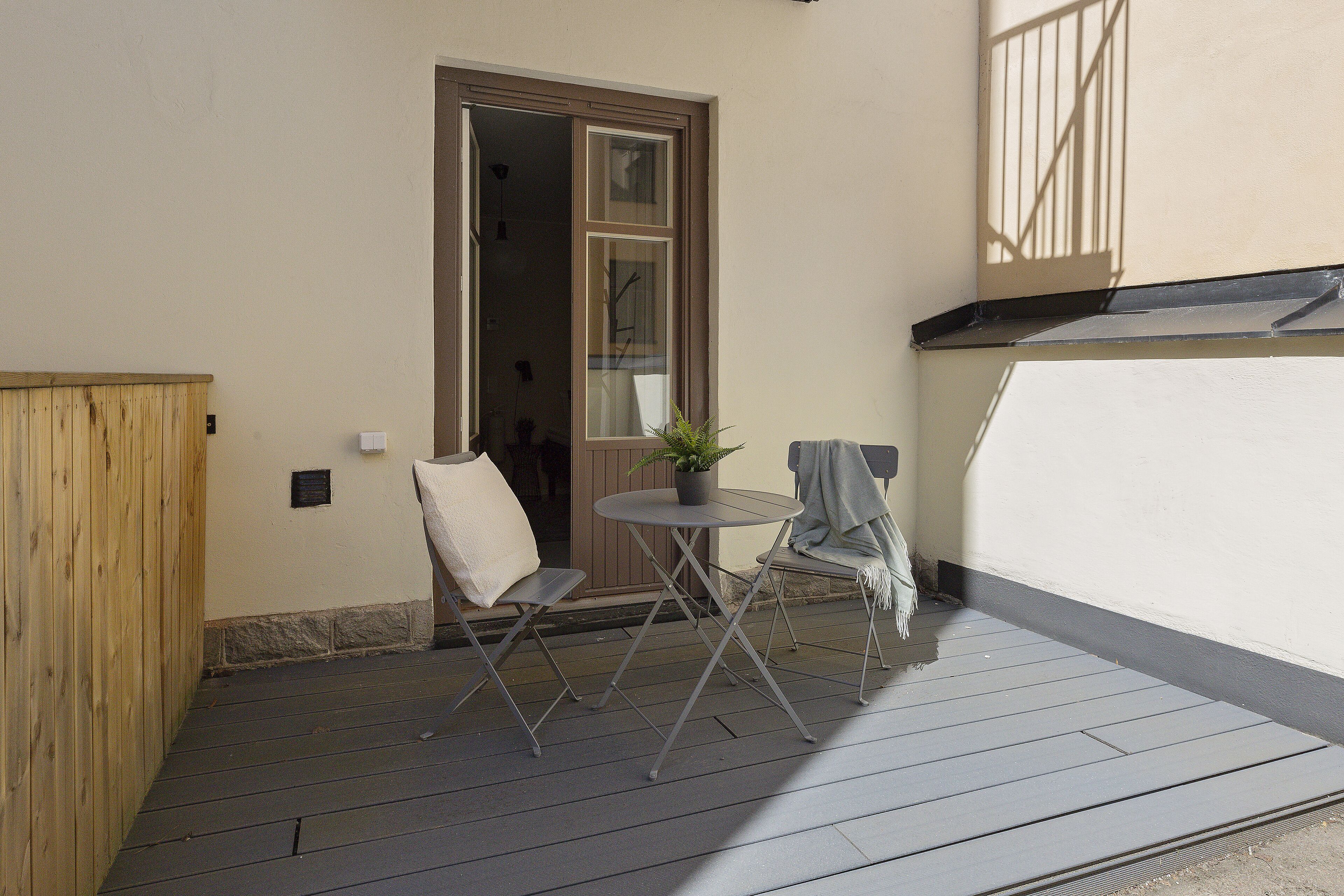 A small outdoor patio with a table and two chairs, a potted plant, and a wooden fence.