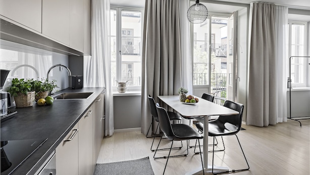 A modern kitchen with a dining area, featuring a large window with curtains, a hanging light fixture, and a dining table with chairs. - Forenom Apartments Stockholm Johannesgatan (Stockholm)