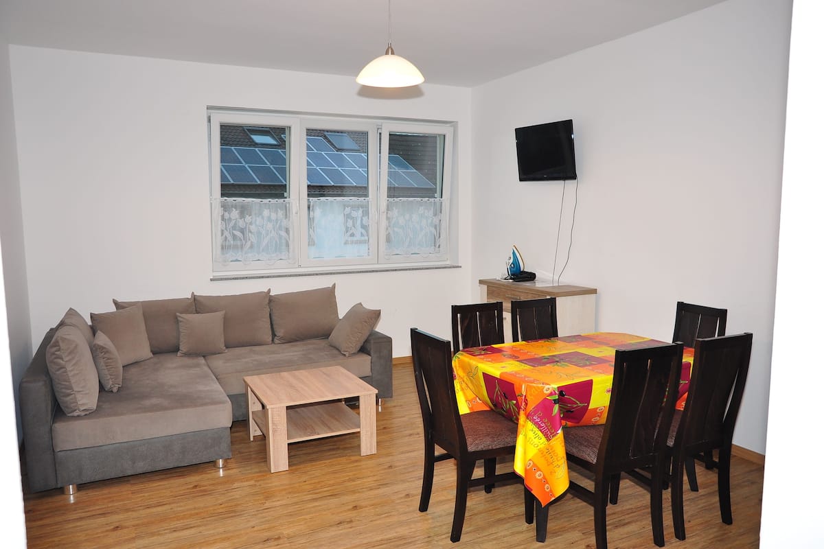 A living room with a gray sofa, wooden coffee table, and a dining area with a table covered in a colorful tablecloth and four chairs.