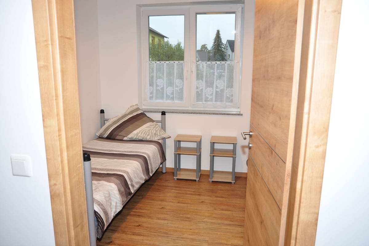 A small bedroom with a bed, two nightstands, a window with a view of houses and trees, and wooden flooring.