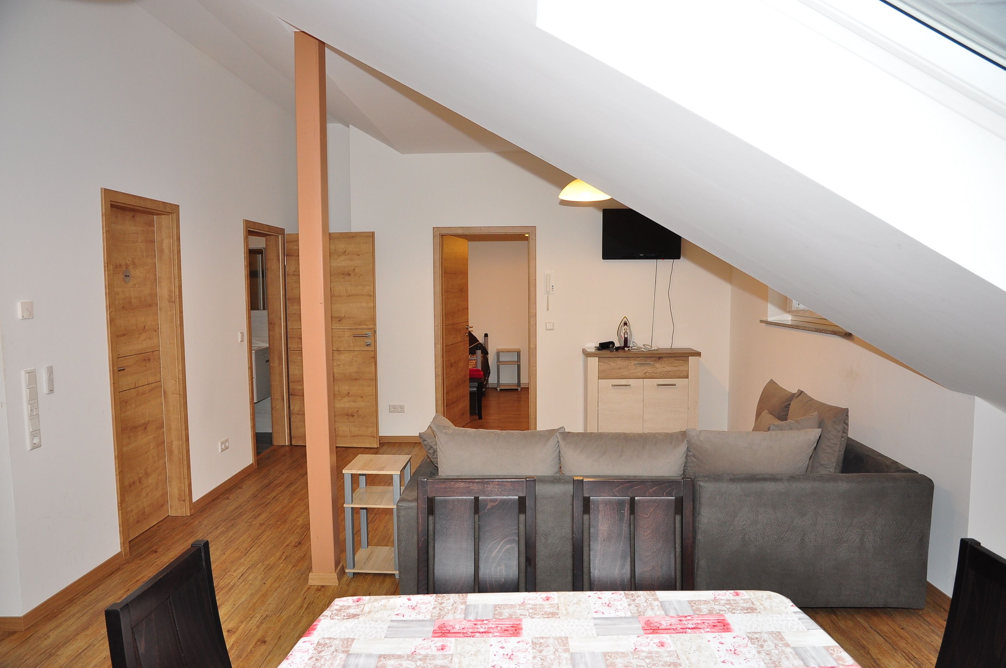 A loft-style apartment with a high ceiling, wooden floors, a sofa, a dining table, and a TV mounted on the wall.