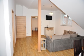 A compact living space with a wooden floor, a grey sofa, a small table, a TV mounted on the wall, and a window with a view. - Ebersheim Luxus Plaza (Mainz)