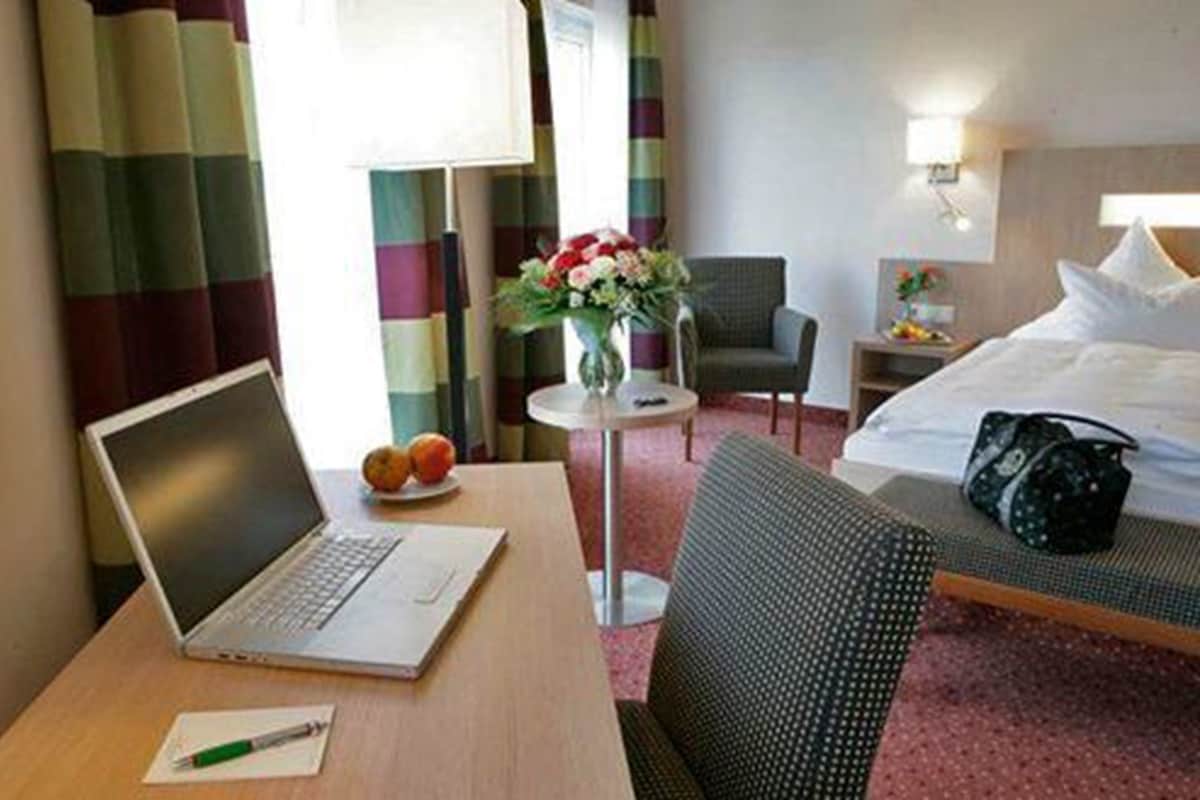 A hotel room with a bed, a desk with a laptop, a chair, a small table with a vase of flowers, and a handbag on the bed.