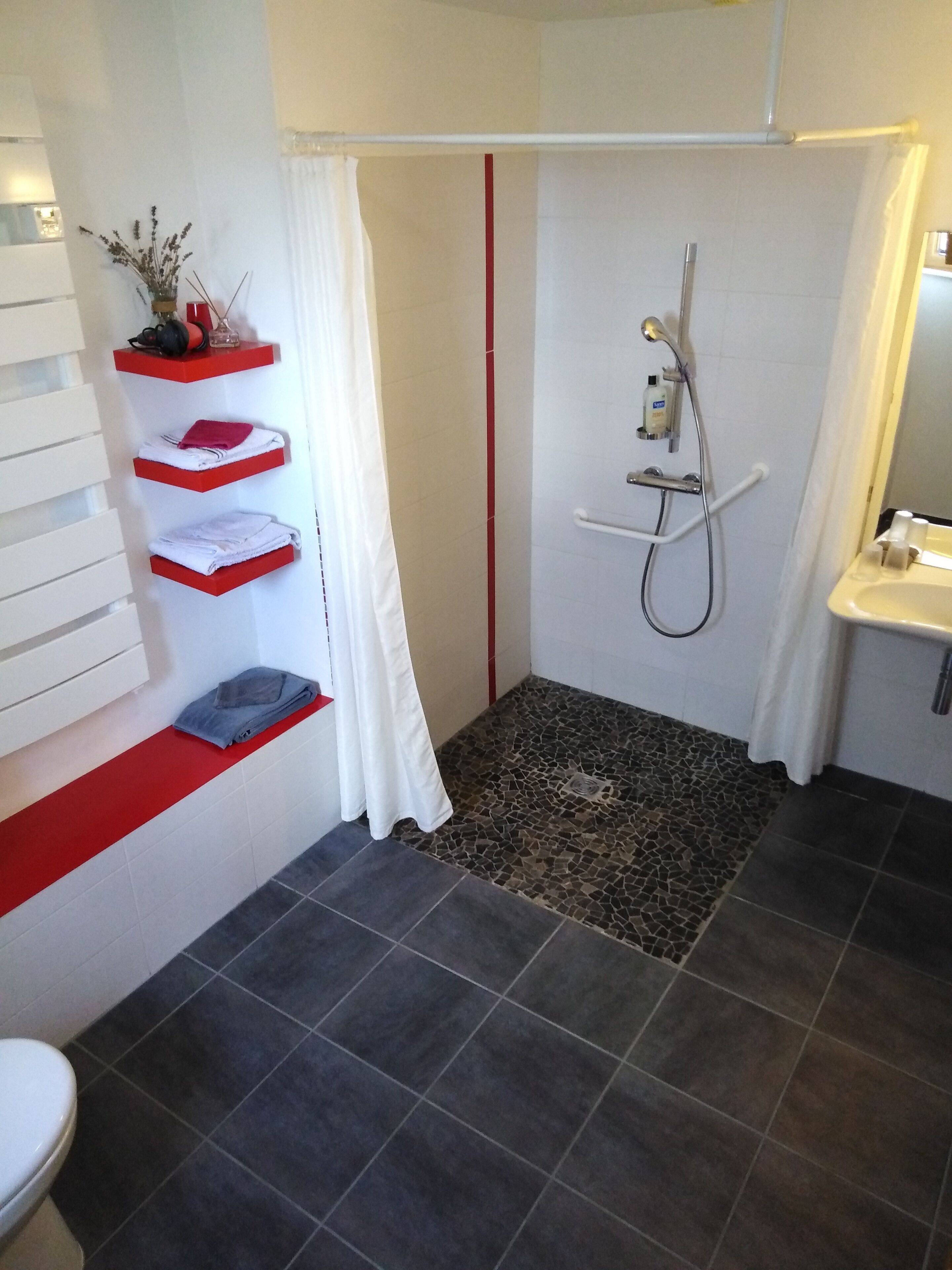 A bathroom with a shower, a toilet, and a sink. There are shelves with towels and toiletries.