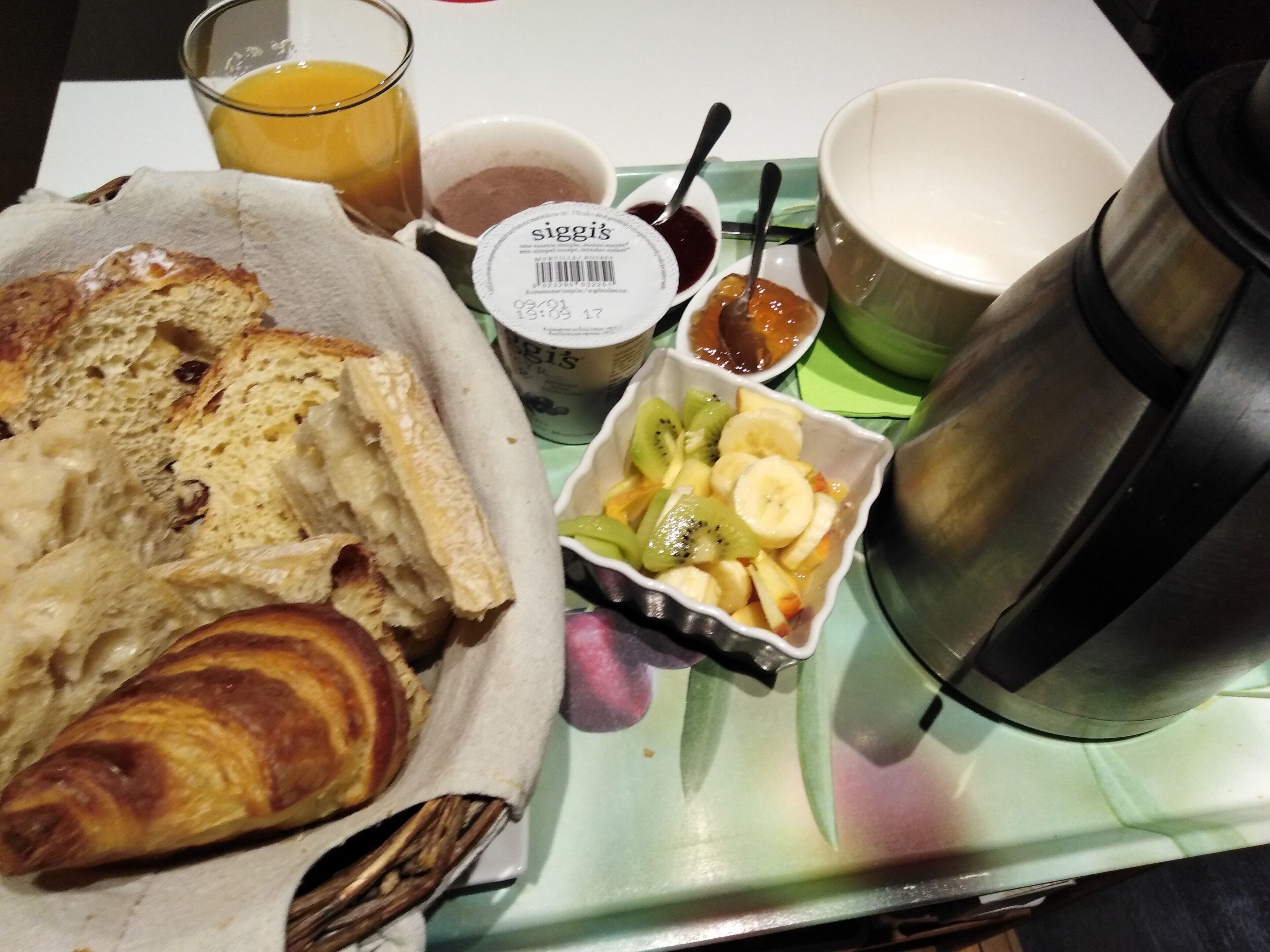 Un plateau de petit-déjeuner avec des croissants, un verre de jus d’orange, un bol de fruits, un pot de yaourt et un thermos.